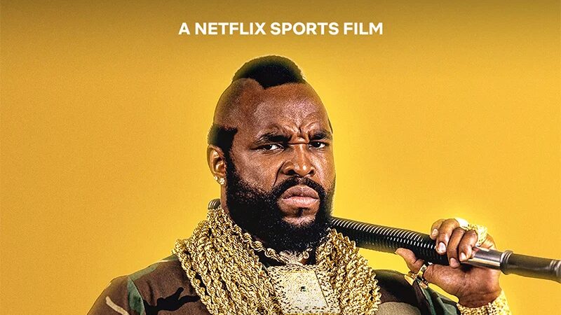 Mr. T stands in camouflage clothing with a mohawk, beard, gold chains and a large championship-style belt while holding a black baton.