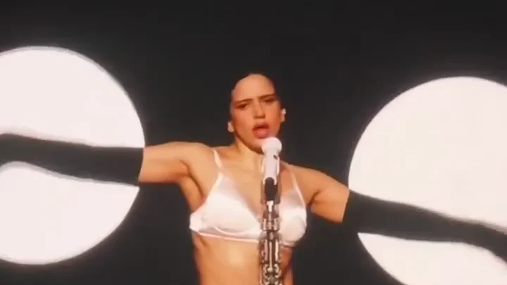 ROSALÍA sings into a microphone with her arms outstretched, surrounded by circular stage lights and a sculptural arrangement of pale arms.