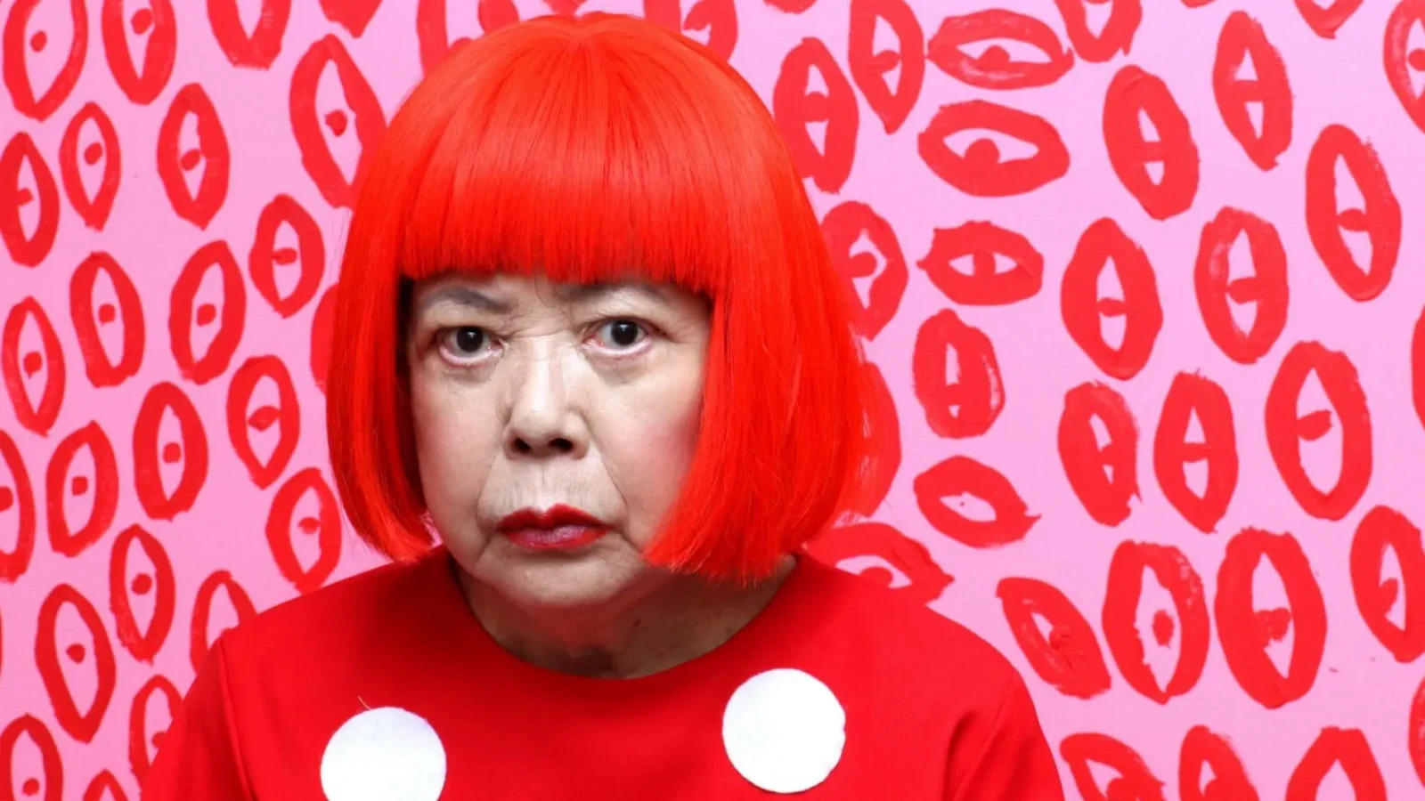 Yayoi Kusama wears a bright red bob wig and red garment with large white dots, posed against a pink wall covered in red abstract motifs.