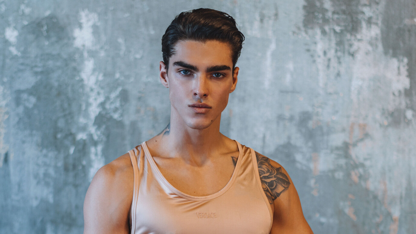 Simone Stravolo looks directly at the camera while wearing a light Versace tank top and black pants against a textured blue-gray wall.
