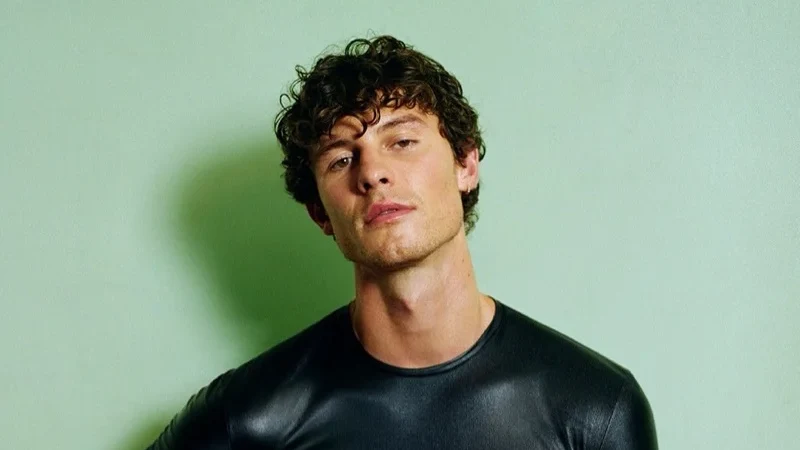 Shawn Mendes poses in a fitted black shirt and black pants against a pale green wall, wearing a black glove and showing tattoos on his arms.