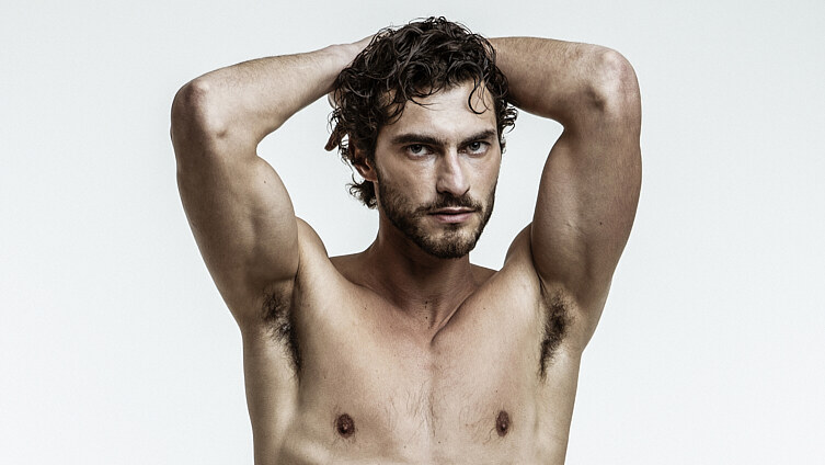 Pedro Pinto poses shirtless with both arms raised, wearing a glossy black wrap skirt against a pale background.