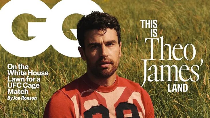 Theo James sits in tall grass wearing a red-and-cream football-style shirt and dark trousers, presented on a magazine cover.