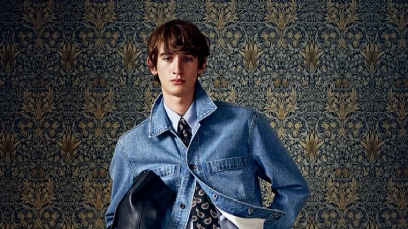 Model wearing a blue denim jacket, patterned necktie, white shirt, light jeans and black loafers while carrying a black leather bag against ornate wallpaper.