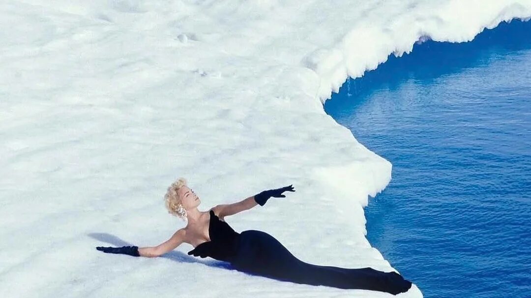 A woman in a black gown and gloves reclines on the edge of a white iceberg above deep blue water.