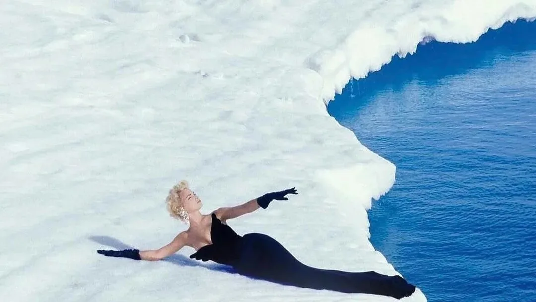 A woman in a black gown and gloves reclines on the edge of a white iceberg above deep blue water.