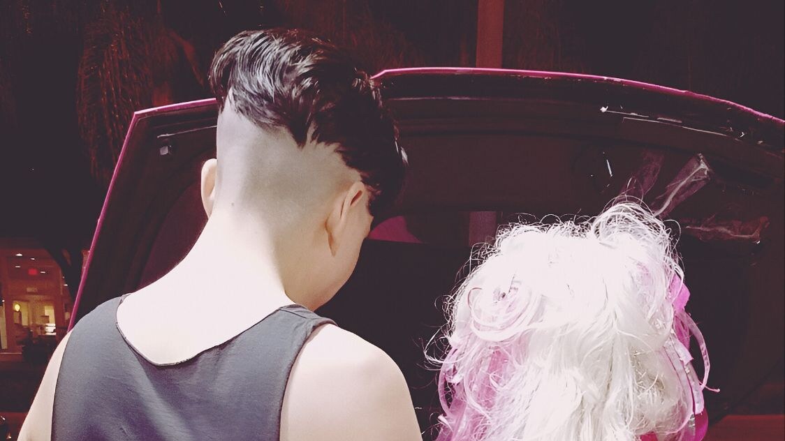 Two people seen from behind stand beside a pink car, one wearing a black sleeveless top and the other in a bright pink jacket with long platinum-blonde hair.