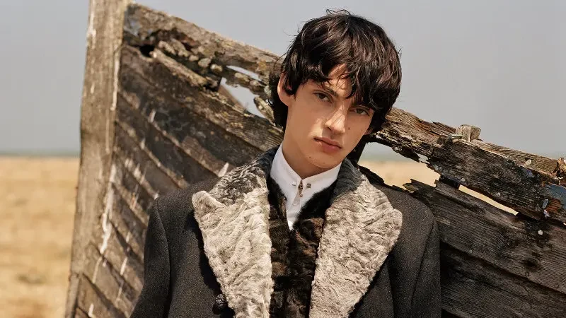 Model wearing a dark tailored coat with a textured fur collar, patterned inner layer and bright red trousers, standing beside weathered wooden fencing in a sandy landscape.