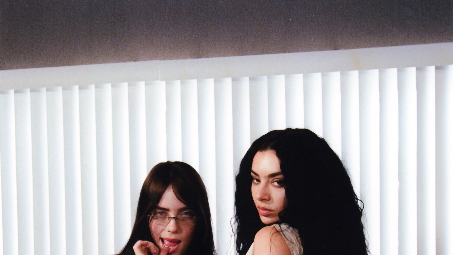 Billie Eilish and Charli xcx pose together in front of white vertical blinds, with Eilish touching her lips and Charli looking toward the camera.