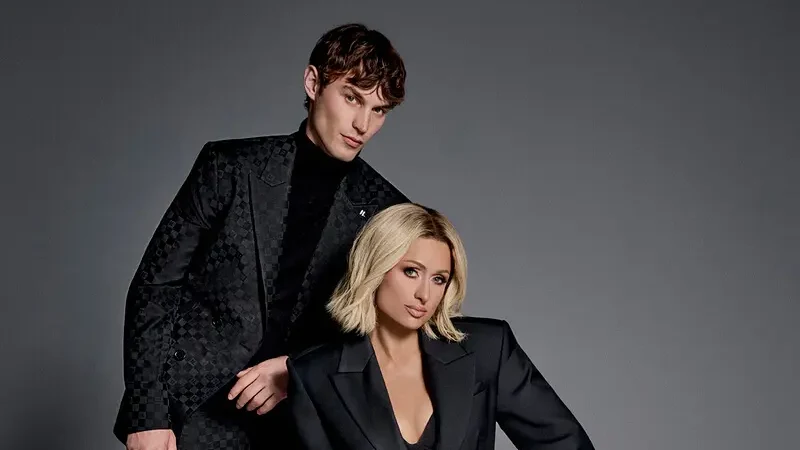 Paris Hilton sits in a black suit beside Kit Butler, with a retro television displaying a monochrome portrait of Karl Lagerfeld.